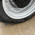 Close-up of a car wheel: black tire with a silver rim, showing the valve stem and lug area on a muddy surface.