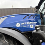 Close-up of a blue New Holland tractor cab hood with vent grills and rain droplets, showing the front lights and part of the cab.