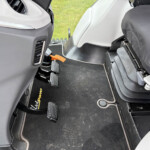 Inside a vehicle cabin showing driver and passenger area with multiple pedals, an orange leash or strap near the pedals, and a black floor mat.