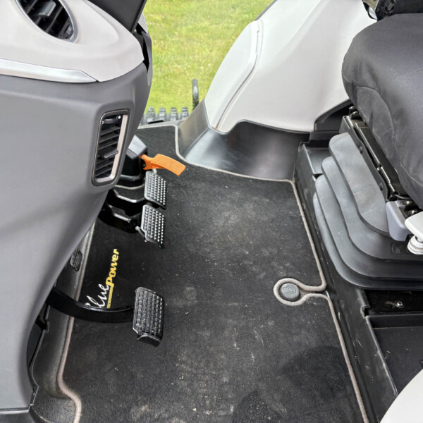 Inside a vehicle cabin showing driver and passenger area with multiple pedals, an orange leash or strap near the pedals, and a black floor mat.