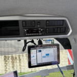 Interior of a vehicle cab with a central dashboard, touchscreen display, and row of switches under a gray overhead panel]**