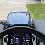 Forward view from inside a blue truck cab showing the steering wheel and digital gauges, with a concrete road and grassy area ahead.