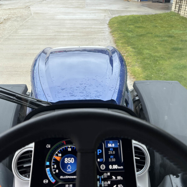 Forward view from inside a blue truck cab showing the steering wheel and digital gauges, with a concrete road and grassy area ahead.