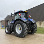 Blue agricultural tractor parked in a yard beside a corrugated metal industrial building, showing oversized rear tires and a yellow license plate on the cab.