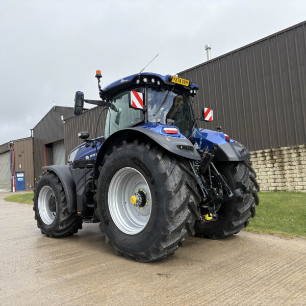 Blue agricultural tractor parked in a yard beside a corrugated metal industrial building, showing oversized rear tires and a yellow license plate on the cab.