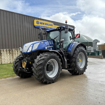 Blue New Holland tractor parked in front of a grey industrial building with a yellow and blue 'New Holland Agriculture' sign above it