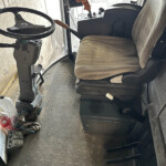 Inside the cab of a New Holland vehicle: steering wheel, orange control lever, and beige driver seat with a dashboard in front.