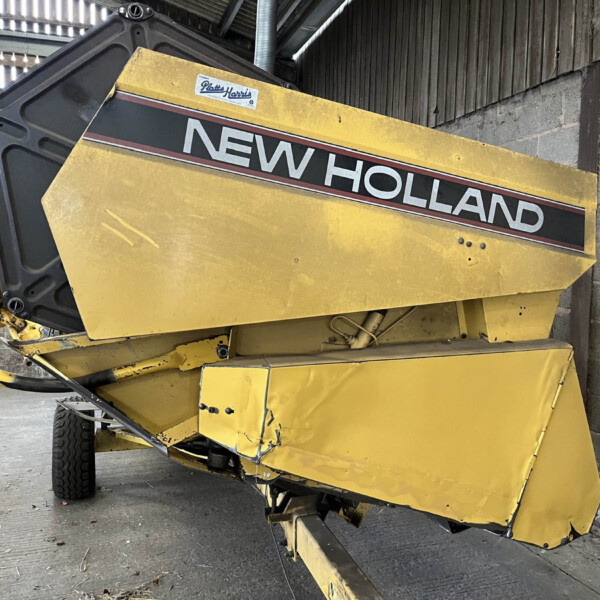 Yellow New Holland agricultural baler inside a workshop, angled with visible hydraulic arms and worn panels.
