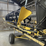 Yellow agricultural harvesting header on a trailer inside a dim workshop, with black frames and metal pegs along the top rail.