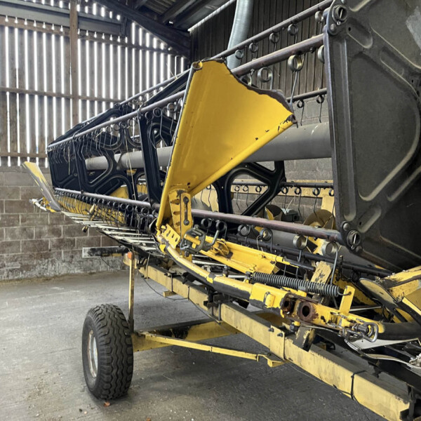 Yellow agricultural harvesting header on a trailer inside a dim workshop, with black frames and metal pegs along the top rail.