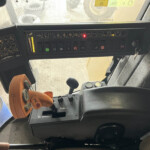 Interior of a tractor cab with a row of switches and levers on the dashboard and a yellow/orange joystick on the left side, illuminated indicators visible near the top, and a view of the field through the windshield.