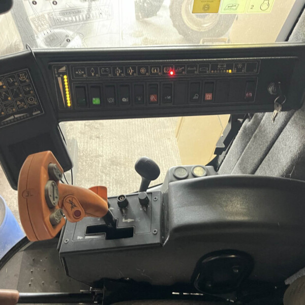Interior of a tractor cab with a row of switches and levers on the dashboard and a yellow/orange joystick on the left side, illuminated indicators visible near the top, and a view of the field through the windshield.