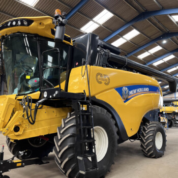 Yellow New Holland combine harvester inside a spacious workshop with blue metal beams and skylights, header raised at the front and large front tires visible.