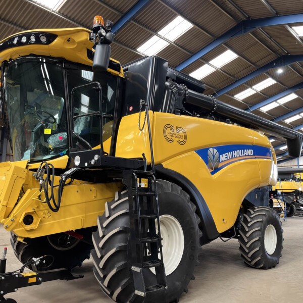 Yellow New Holland combine harvester inside a spacious workshop with blue metal beams and skylights, header raised at the front and large front tires visible.