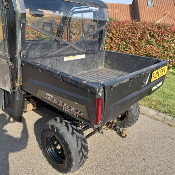 Black Polaris Ranger Diesel utility vehicle with a large flatbed, parked on a driveway near a hedge and red-tiled roof houses behind it.