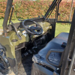 Interior view of a forklift cabin showing steering wheel, controls, and a torn seat with taped repairs outside a hedge backdrop