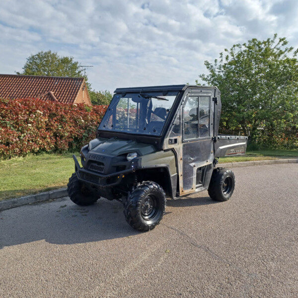 Gray Polaris Ranger utility vehicle parked along a curb in a suburban yard, with red hedges and trees under a cloudy sky