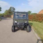 Front view of a dark gray Polaris off-road utility vehicle parked on a suburban street with a hedge and brick house in the background
