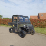 Black utility side-by-side ATV with a roll cage parked on a residential street, red hedge and red-tiled house in the background.