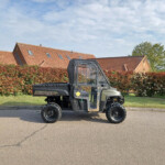 Off-road utility vehicle (Ranger Diesel) parked on a street with red-tiled-roof houses and a green hedge behind it.