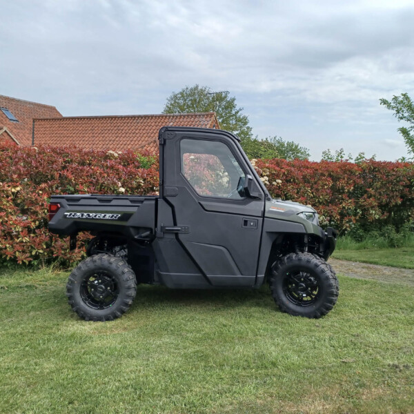 Front-side view of a black Ranger off-road utility vehicle parked on a grassy lawn, with a red-leaved hedge and a red-tiled roof house in the background.