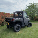Black Polaris Ranger off-road utility vehicle parked on a grassy lawn beside red-leaved shrubs and green trees under a cloudy sky.