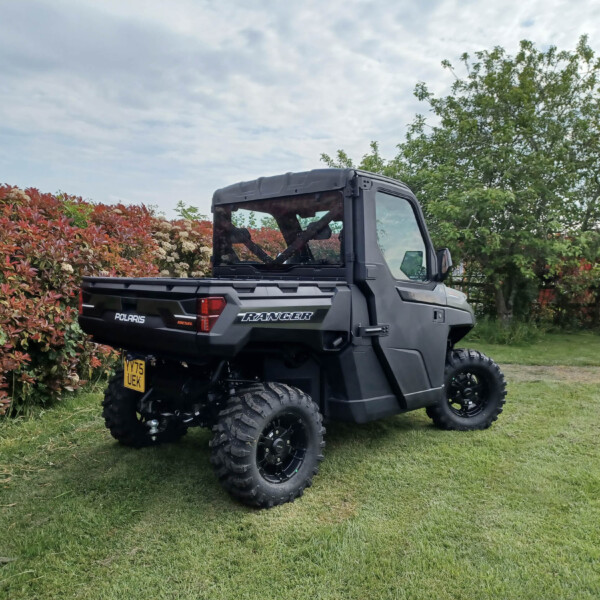 Black Polaris Ranger off-road utility vehicle parked on a grassy lawn beside red-leaved shrubs and green trees under a cloudy sky.