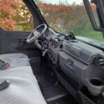 Interior of a pickup truck cab with a gray fabric seat, steering wheel, and dashboard; hedge visible through the windows outside.