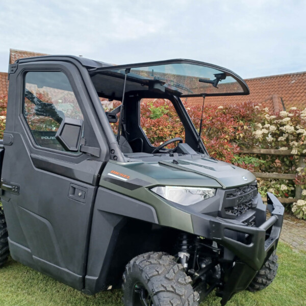 Close-up of a dark Polaris Ranger off-road utility vehicle with open cabin, parked on grass with red-leaved shrubs and a brick house in the background.