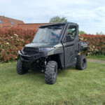 Gray Polaris Ranger UTV parked on a grassy lawn beside a red-leaf hedge and a tiled-roof house in the background, under a cloudy sky.