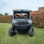 Front view of a Polaris off-road vehicle with an open cabin, parked on a grassy lawn with red hedges and a brick-roof house in the background.
