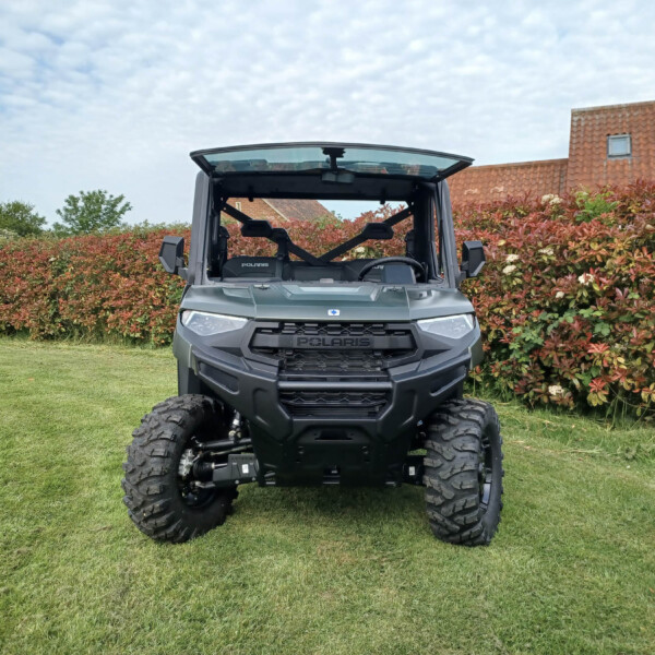 Front view of a Polaris off-road vehicle with an open cabin, parked on a grassy lawn with red hedges and a brick-roof house in the background.