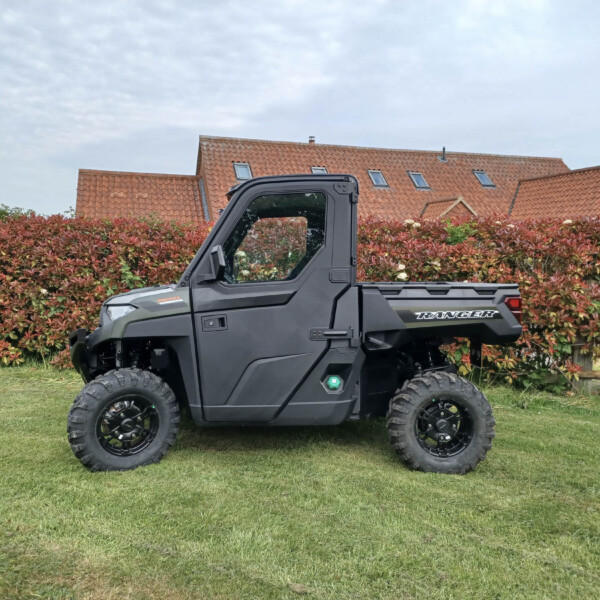 Black utility off-road vehicle (Ranger) parked on a grassy lawn with a red hedge and red-tiled houses in the background.