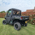 Polaris Ranger off-road utility vehicle parked on a grassy lawn beside a red-leaf hedge and a tiled roof house nearby.