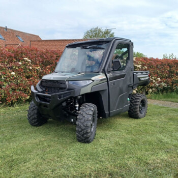 Gray Polaris Ranger UTV parked on a grassy lawn beside a red-leaf hedge and a tiled-roof house in the background, under a cloudy sky.