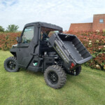 Black off-road utility vehicle with a raised dump bed on a grassy lawn, with red-leaved shrubs and a house with a tiled roof in the background.