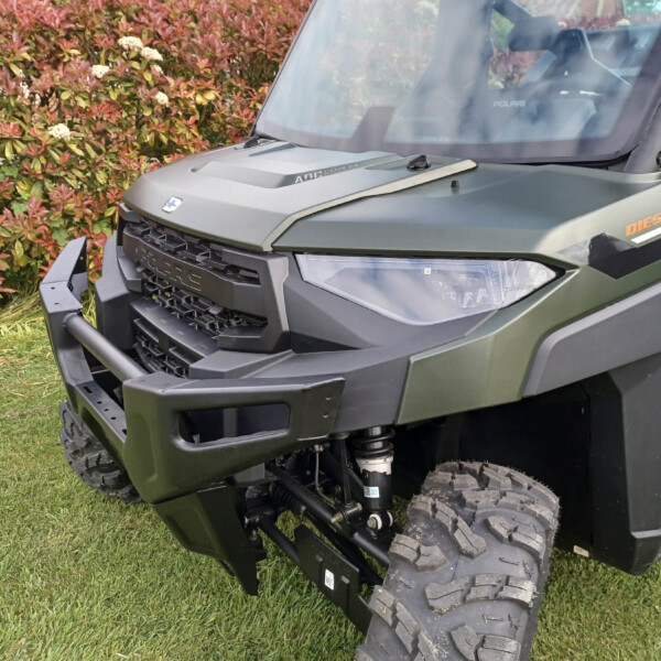 Close-up of a rugged off-road vehicle front ('Polaris' branding) with a diesel badge, green matte body, and large treaded tires, outdoors by colorful shrubs.