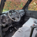 Interior view of a rugged off-road vehicle cockpit with steering wheel, dashboard, pedals, and cup holder, seats covered in light gray upholstery; colorful shrubbery visible through the windows.