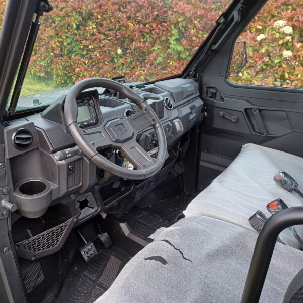 Interior view of a rugged off-road vehicle cockpit with steering wheel, dashboard, pedals, and cup holder, seats covered in light gray upholstery; colorful shrubbery visible through the windows.