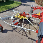 Red and yellow agricultural implement with multiple tines parked on a suburban driveway/road edge on a sunny day.