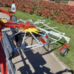 Red and white agricultural equipment with folding arms and wheels resting on a driveway beside a hedge.