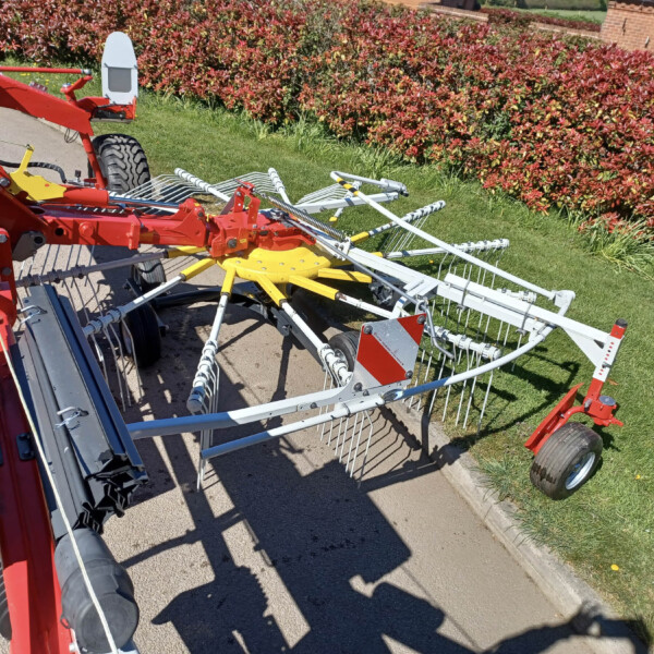 Red and white agricultural equipment with folding arms and wheels resting on a driveway beside a hedge.