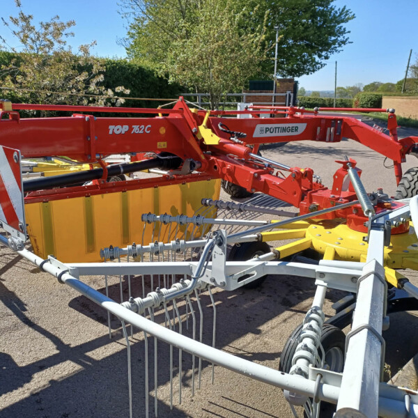 Red Pottinger TOP 762C agricultural implement with yellow frame and multiple tines, parked outdoors near hedges and trees.