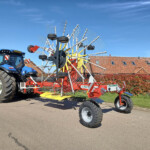 Blue tractor pulling a large red hay rake with many white tines outdoors on a sunny day, with houses in the background.