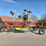 Red agricultural rotary rake with multiple spoke arms and tines, parked in a driveway under a blue sky.
