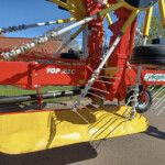 Close-up of a red and yellow agricultural header with hydraulic linkages and rotating tines, mounted on a trailer in a sunny outdoor setting.