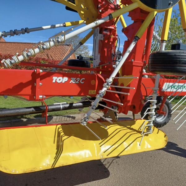 Close-up of a red and yellow agricultural header with hydraulic linkages and rotating tines, mounted on a trailer in a sunny outdoor setting.