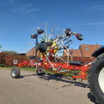 Large red farm implement with multiple metal tines and wheels, parked in a lot under a clear blue sky.