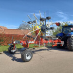 Blue tractor pulling a large red multi-row hay rake with metal tines on a sunny day.