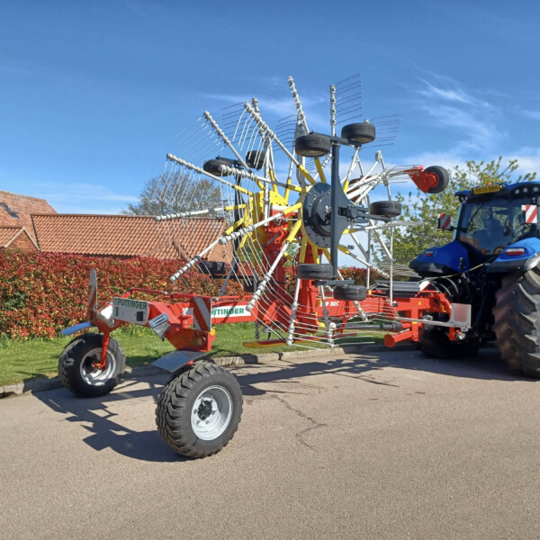 Blue tractor pulling a large red multi-row hay rake with metal tines on a sunny day.